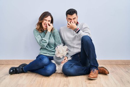 Photo for Young hispanic couple sitting on the floor with dog smelling something stinky and disgusting, intolerable smell, holding breath with fingers on nose. bad smell - Royalty Free Image