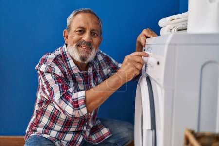 Téléchargez les photos : Senior homme aux cheveux gris souriant confiant tournant sur la machine à laver à la buanderie - en image libre de droit