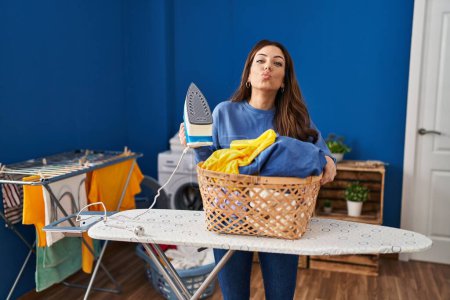 Photo for Young brunette woman ironing clothes at home looking at the camera blowing a kiss being lovely and sexy. love expression. - Royalty Free Image