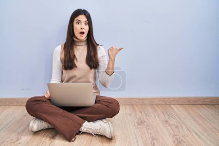 Foto de Mujer morena joven trabajando con ordenador portátil sentado en el suelo sorprendido señalando con el dedo de la mano a un lado, boca abierta expresión asombrada. - Imagen libre de derechos