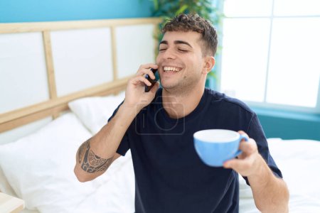 Foto de Hombre hispano joven hablando en smartphone tomando café en el dormitorio - Imagen libre de derechos