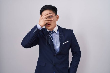 Foto de Joven hombre asiático vistiendo traje de negocios y corbata espiando en shock cubriendo la cara y los ojos con la mano, mirando a través de los dedos con expresión avergonzada. - Imagen libre de derechos