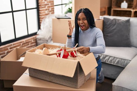 Téléchargez les photos : African american woman moving to a new home opening boxes celebrating victory with happy smile and winner expression with raised hands - en image libre de droit