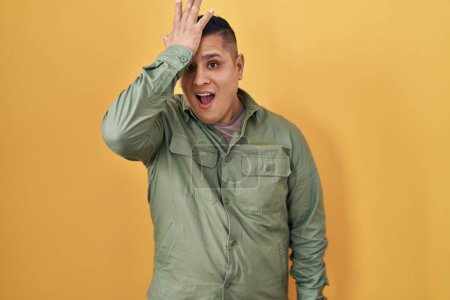 Foto de Hispanic young man standing over yellow background surprised with hand on head for mistake, remember error. forgot, bad memory concept. - Imagen libre de derechos