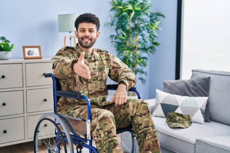Foto de Hombre árabe vistiendo camuflaje uniforme del ejército sentado en silla de ruedas sonriendo amistoso ofreciendo apretón de manos como saludo y bienvenida. negocios exitosos. - Imagen libre de derechos