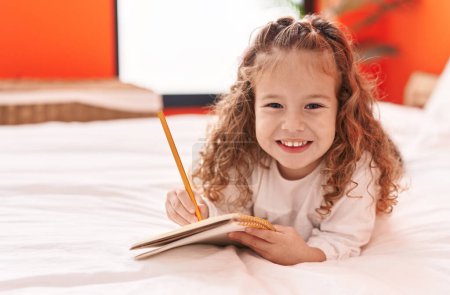 Photo for Adorable blonde toddler writing on notebook lying on bed at bedroom - Royalty Free Image