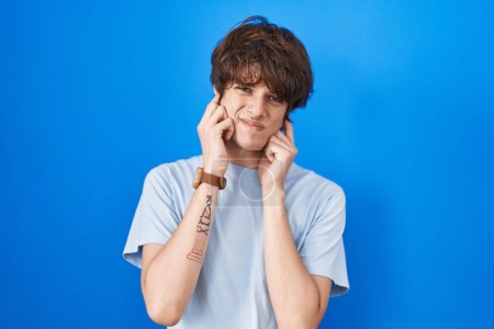 Photo for Hispanic young man standing over blue background covering ears with fingers with annoyed expression for the noise of loud music. deaf concept. - Royalty Free Image
