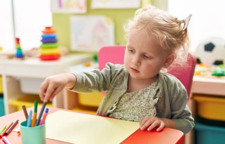Foto de Adorable blonde girl preschool student sitting on table drawing on paper at kindergarten - Imagen libre de derechos