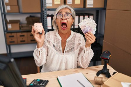 Photo for Middle age woman with grey hair working at small business ecommerce holding piggy bank and zloty angry and mad screaming frustrated and furious, shouting with anger looking up. - Royalty Free Image
