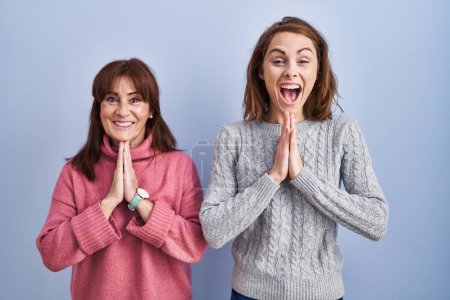 Photo for Mother and daughter standing over blue background praying with hands together asking for forgiveness smiling confident. - Royalty Free Image