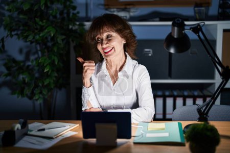 Foto de Middle age woman working at the office at night smiling with happy face looking and pointing to the side with thumb up. - Imagen libre de derechos