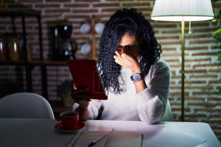 Foto de Middle age hispanic woman using touchpad sitting on the table at night tired rubbing nose and eyes feeling fatigue and headache. stress and frustration concept. - Imagen libre de derechos