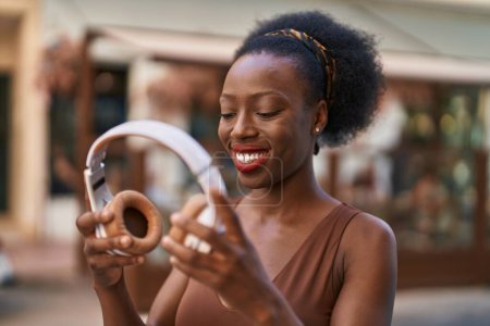 Téléchargez les photos : Afro-américaine femme souriant confiant tenant écouteurs à la rue - en image libre de droit
