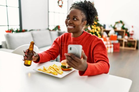 Téléchargez les photos : African american woman eating hamburger using smartphone sitting on table by christmas tree at home - en image libre de droit