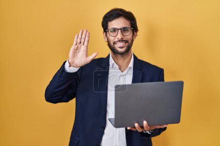 Foto de Hombre latino guapo trabajando usando computadora portátil renunciando a decir hola feliz y sonriente, gesto de bienvenida amistoso - Imagen libre de derechos