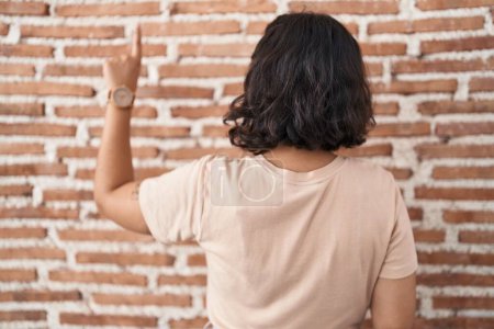 Foto de Mujer hispana joven de pie sobre la pared de ladrillos posando hacia atrás apuntando hacia adelante con la mano del dedo - Imagen libre de derechos