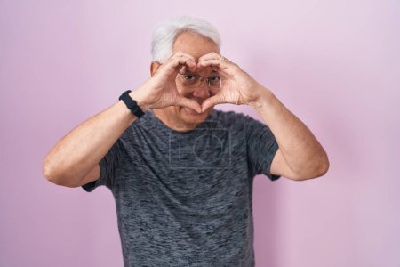 Téléchargez les photos : Middle age man with grey hair standing over pink background doing heart shape with hand and fingers smiling looking through sign - en image libre de droit