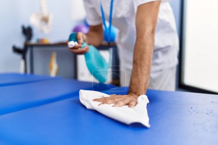 Téléchargez les photos : Young hispanic man wearing physiotherapist uniform disinfecting massage table at rehab clinic - en image libre de droit
