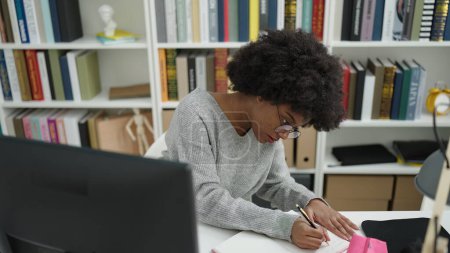 Téléchargez les photos : African american woman student using computer writing on notebook at library university - en image libre de droit