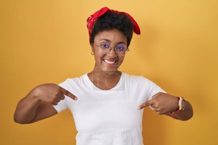 Foto de Joven mujer afroamericana de pie sobre fondo amarillo mirando confiado con sonrisa en la cara, señalándose con los dedos orgullosos y felices. - Imagen libre de derechos