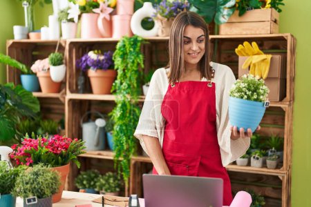 Photo for Young beautiful hispanic woman florist using laptop holding plant at flower shop - Royalty Free Image