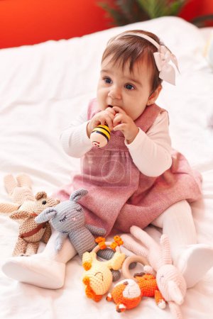 Foto de Adorable bebé hispano mordiendo maraca sentado en la cama en el dormitorio - Imagen libre de derechos