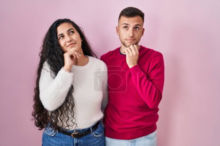 Foto de Young hispanic couple standing over pink background with hand on chin thinking about question, pensive expression. smiling with thoughtful face. doubt concept. - Imagen libre de derechos