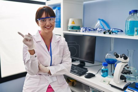 Téléchargez les photos : Young brunette woman working at scientist laboratory with a big smile on face, pointing with hand and finger to the side looking at the camera. - en image libre de droit