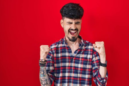 Téléchargez les photos : Jeune homme hispanique avec la barbe debout sur fond rouge excité pour le succès avec les bras levés et les yeux fermés célébrant la victoire en souriant. concept gagnant. - en image libre de droit