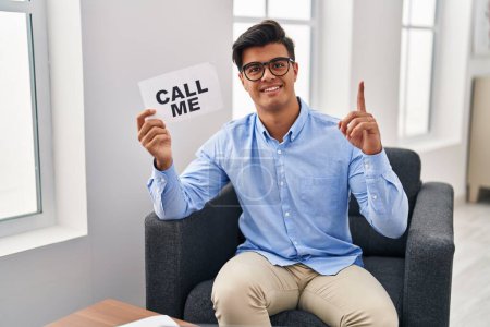 Foto de Hispanic man working at therapy office holding call me banner surprised with an idea or question pointing finger with happy face, number one - Imagen libre de derechos