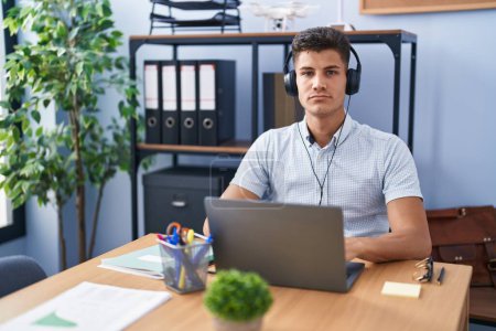 Téléchargez les photos : Young hispanic man working at the office wearing headphones relaxed with serious expression on face. simple and natural looking at the camera. - en image libre de droit