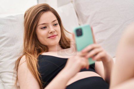 Téléchargez les photos : Jeune femme caucasienne utilisant smartphone couché sur le lit dans la chambre - en image libre de droit