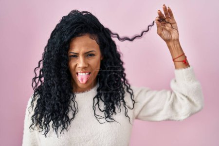 Foto de Mujer hispana de mediana edad sosteniendo el pelo rizado sacando la lengua feliz con expresión divertida. - Imagen libre de derechos