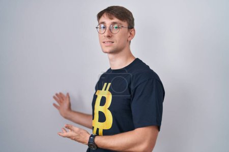 Foto de Hombre rubio caucásico con camiseta bitcoin invitando a entrar sonriendo natural con la mano abierta - Imagen libre de derechos
