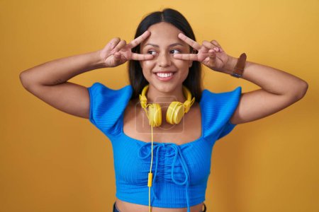 Téléchargez les photos : Hispanic young woman standing over yellow background doing peace symbol with fingers over face, smiling cheerful showing victory - en image libre de droit