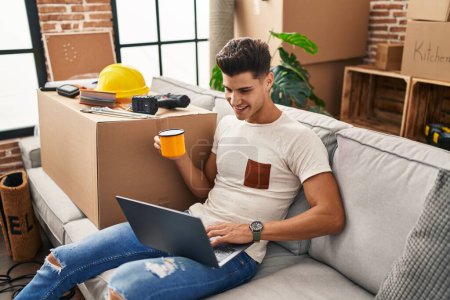 Photo for Young hispanic man using laptop and drinking coffee sitting on sofa at new home - Royalty Free Image