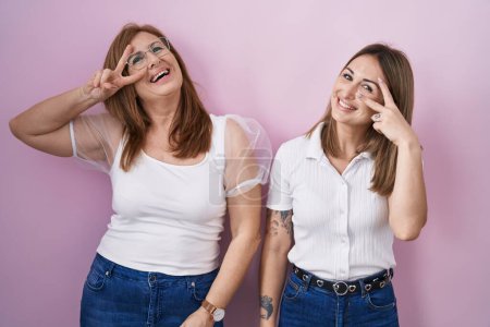Foto de Madre e hija hispanas vistiendo camiseta blanca casual sobre fondo rosa haciendo símbolo de paz con los dedos sobre la cara, sonriendo alegre mostrando la victoria - Imagen libre de derechos