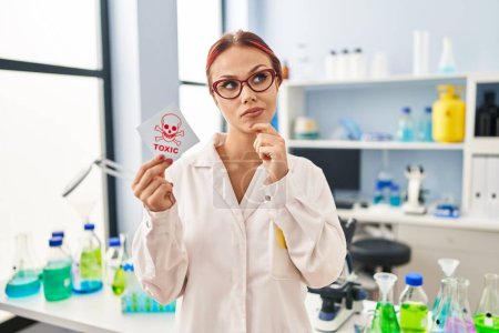 Foto de Young caucasian woman working at scientist laboratory holding toxic label serious face thinking about question with hand on chin, thoughtful about confusing idea - Imagen libre de derechos