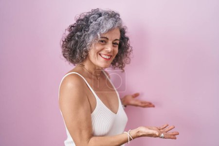 Foto de Middle age woman with grey hair standing over pink background inviting to enter smiling natural with open hand - Imagen libre de derechos