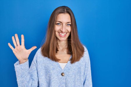 Foto de Mujer joven de pie sobre fondo azul mostrando y señalando hacia arriba con los dedos número cinco mientras sonríe confiado y feliz. - Imagen libre de derechos