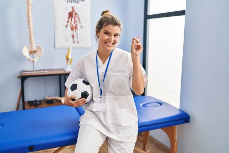 Téléchargez les photos : Jeune femme travaillant à la clinique de thérapie de football souriant heureux pointant avec la main et le doigt vers le côté - en image libre de droit