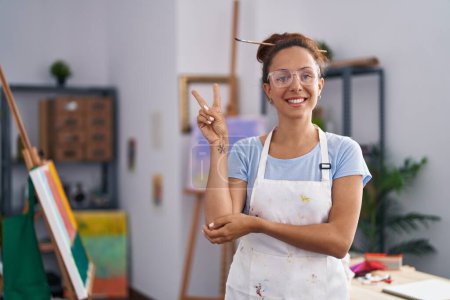 Photo for Brunette woman painting at art studio smiling with happy face winking at the camera doing victory sign. number two. - Royalty Free Image