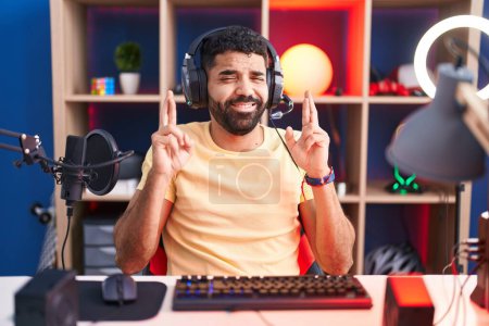 Téléchargez les photos : Hispanic man with beard playing video games with headphones gesturing finger crossed smiling with hope and eyes closed. luck and superstitious concept. - en image libre de droit