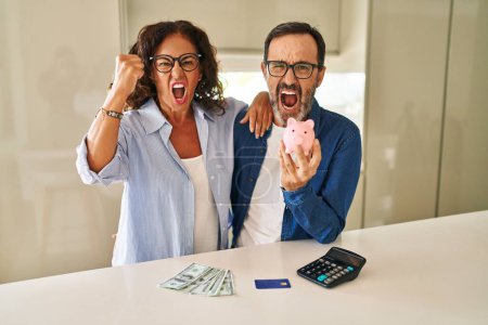 Téléchargez les photos : Middle age couple holding piggy bank calculating savings annoyed and frustrated shouting with anger, yelling crazy with anger and hand raised - en image libre de droit