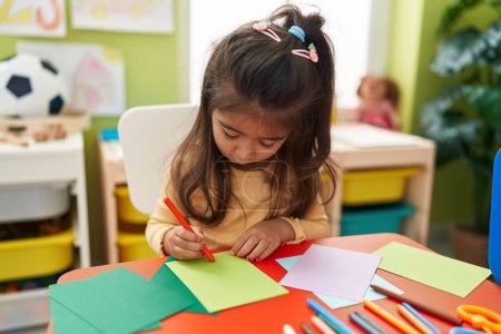 Foto de Adorable hispanic girl preschool student sitting on table drawing on paper at kindergarten - Imagen libre de derechos