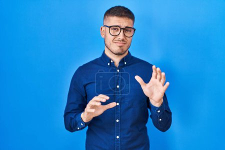 Téléchargez les photos : Young hispanic man wearing glasses over blue background disgusted expression, displeased and fearful doing disgust face because aversion reaction. with hands raised - en image libre de droit