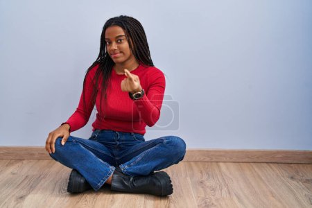 Foto de Young african american with braids sitting on the floor at home beckoning come here gesture with hand inviting welcoming happy and smiling - Imagen libre de derechos