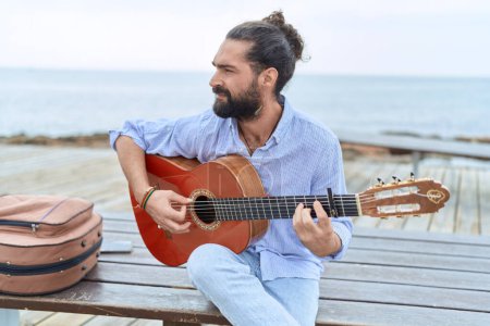 Téléchargez les photos : Jeune homme hispanique musicien jouant de la guitare classique assis sur un banc au bord de la mer - en image libre de droit