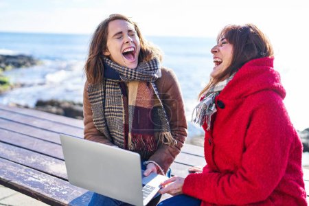 Foto de Two women mother and daughter using laptop sitting on bench at seaside - Imagen libre de derechos