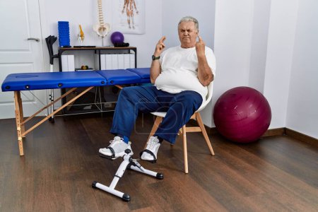 Photo for Senior caucasian man at physiotherapy clinic using pedal exerciser gesturing finger crossed smiling with hope and eyes closed. luck and superstitious concept. - Royalty Free Image